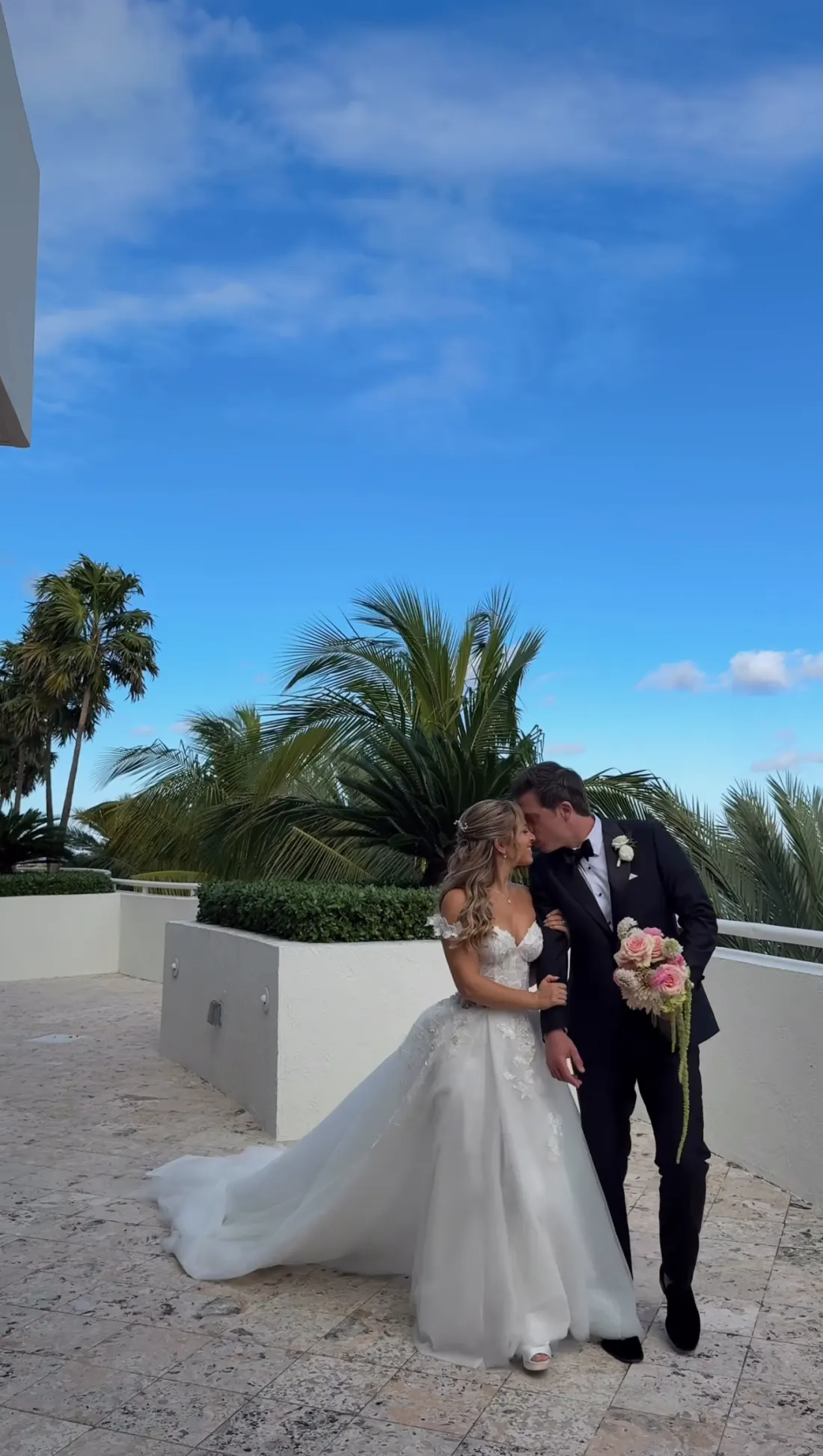 Groom kissing bride's forehead on a sunlit terrace under a wide blue sky