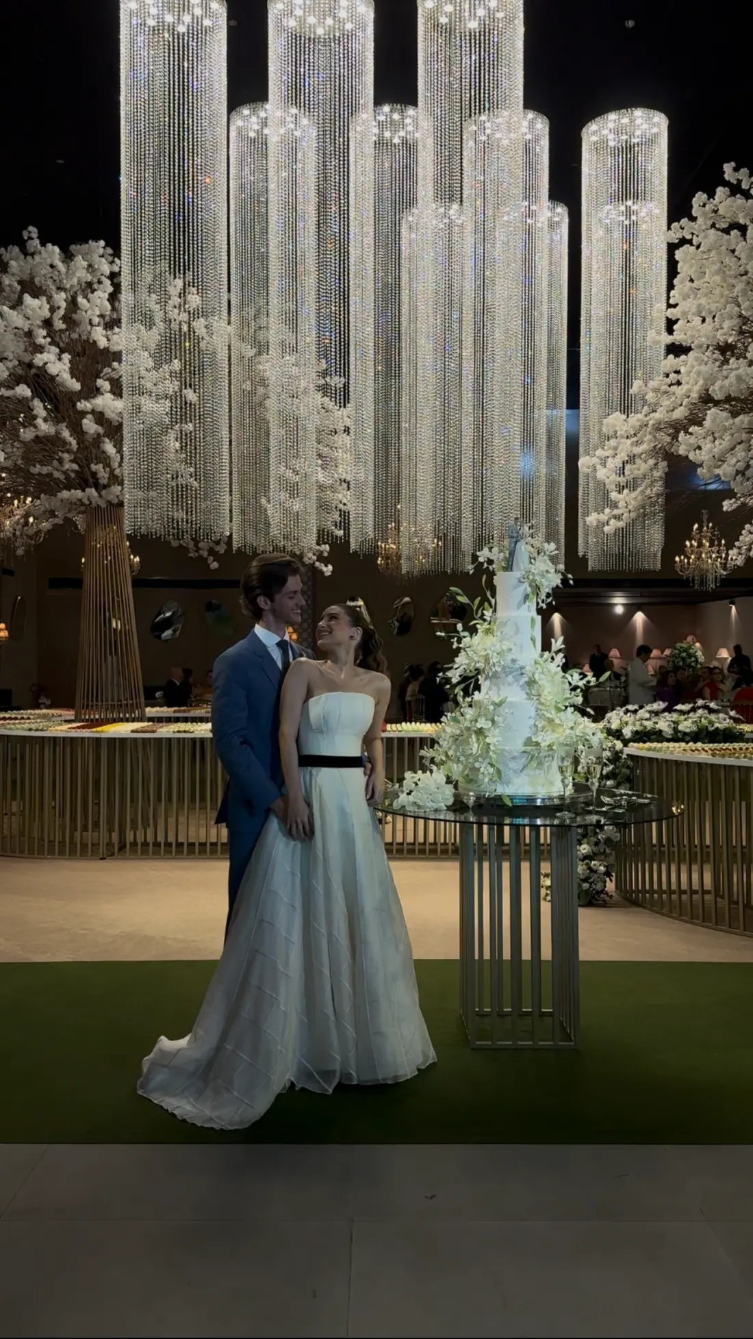 Couple beside a tiered cake beneath towering crystal chandeliers
