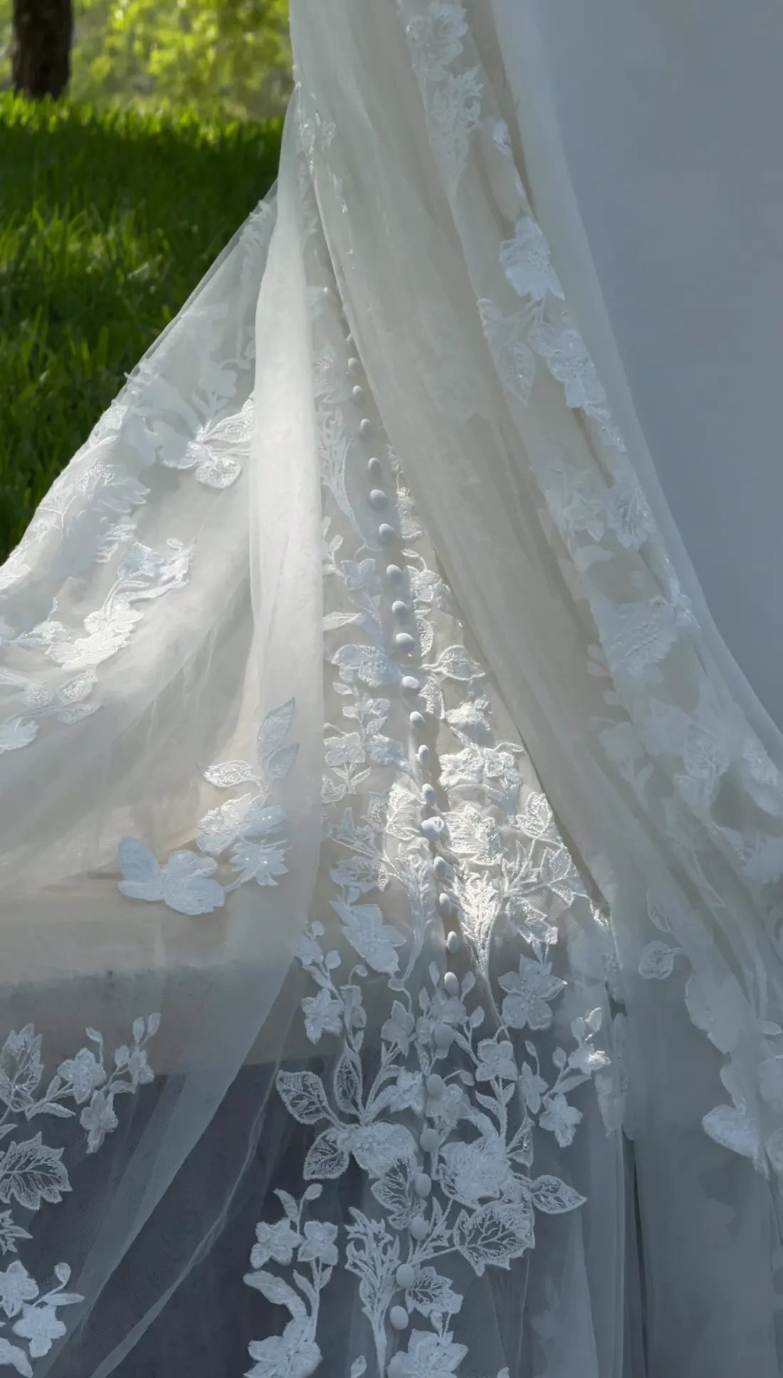 Sunlit lace wedding train with covered buttons against green lawn
